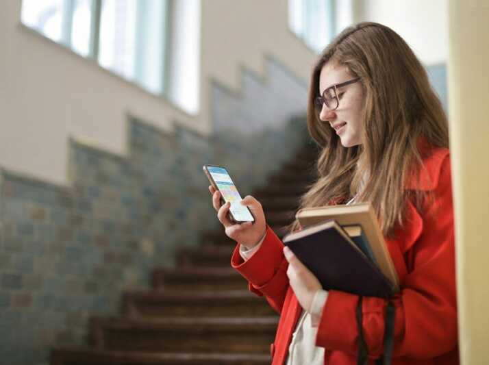 Étudiante portant des lunettes et un manteau rouge, tenant des livres et consultant son smartphone dans un escalier.