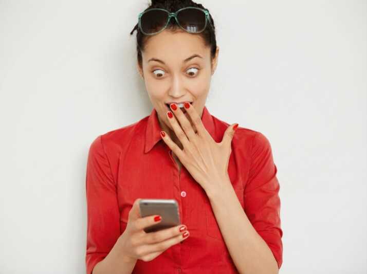 Femme portant une chemise rouge et des lunettes de soleil, regardant son téléphone avec la main devant la bouche.