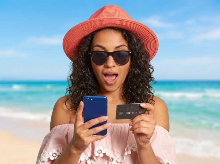 Femme avec chapeau, lunettes, smartphone et carte bancaire sur fond de plage.