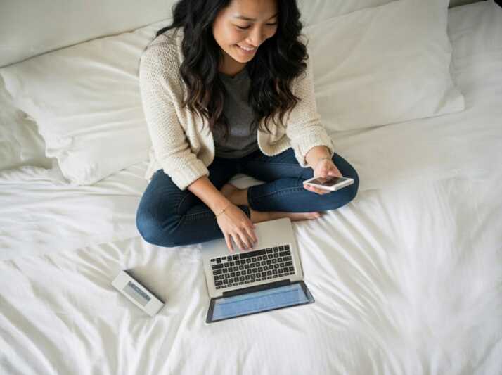 Une femme assise sur un lit utilise un ordinateur portable et un smartphone.