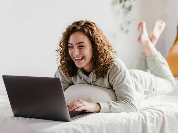 Une jeune femme aux cheveux bouclés allongée sur un lit utilise un ordinateur portable.