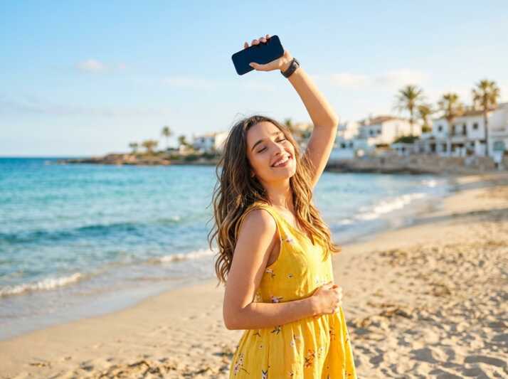 Femme en robe jaune tenant un smartphone levé sur une plage avec la mer en arrière-plan.
