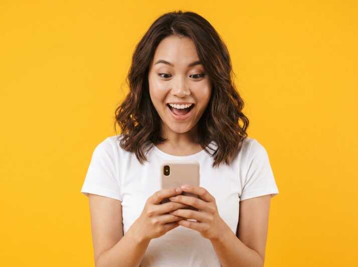 Jeune femme regardant son smartphone avec la bouche ouverte sur fond jaune.
