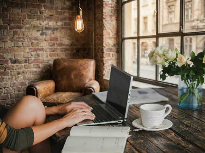 Personne travaillant sur un ordinateur portable à une table en bois avec un carnet, une tasse et des fleurs.