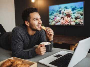Une femme allongée sur un canapé avec un ordinateur portable, une tasse et une télévision affichant un récif corallien.