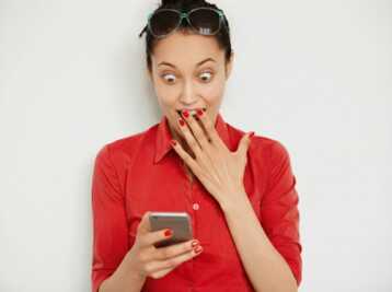 Femme portant une chemise rouge et des lunettes de soleil, regardant son téléphone avec la main devant la bouche.