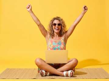Femme en tenue d'été assise sur un tapis avec un ordinateur portable, bras levés sur fond jaune.