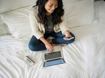 Une femme assise sur un lit utilise un ordinateur portable et un smartphone.