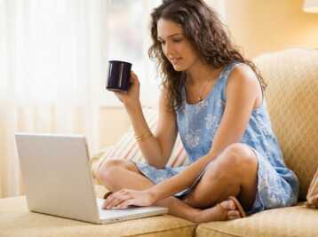 Femme assise sur un canapé, portant une robe bleue, utilisant un ordinateur portable et tenant une tasse.