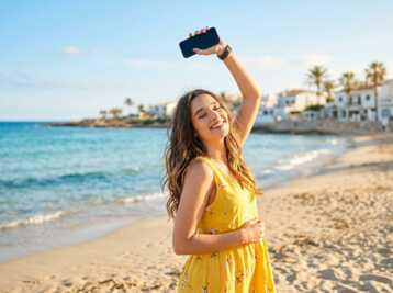 Femme en robe jaune tenant un smartphone levé sur une plage avec la mer en arrière-plan.