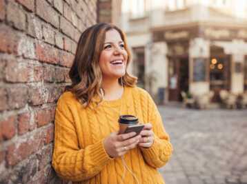 Femme en pull jaune avec un gobelet et un smartphone, adossée à un mur en briques dans une rue.