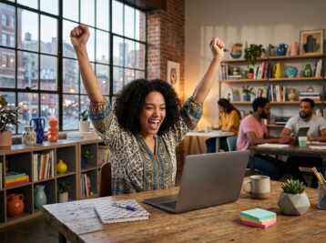 Une femme aux cheveux bouclés lève les bras en signe de victoire devant son ordinateur dans un bureau.