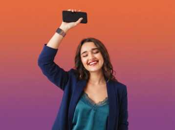 Une femme souriante tenant un smartphone en l'air sur un fond dégradé orange et violet.