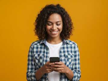 Femme aux cheveux bouclés portant une chemise à carreaux, regardant son smartphone sur fond jaune.