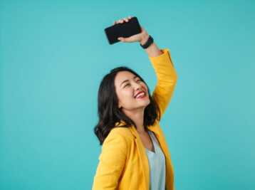 Une femme sourit en levant un smartphone au-dessus de sa tête sur un fond uni.