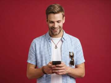 Homme souriant portant une chemise à carreaux et un collier boussole, regardant son téléphone sur fond rouge.