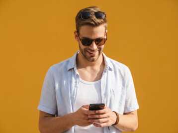 Homme portant des lunettes de soleil et une chemise, utilisant un smartphone devant un mur jaune.
