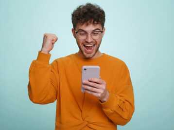 Homme portant un pull orange et des lunettes, tenant un smartphone et levant le poing sur fond bleu.