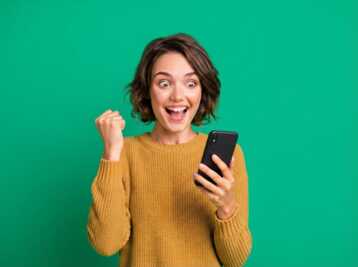 Une personne portant un pull jaune regarde un smartphone avec le poing levé sur fond vert.