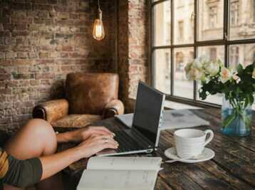 Personne travaillant sur un ordinateur portable à une table en bois avec un carnet, une tasse et des fleurs.