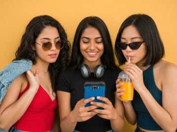 Trois jeunes femmes regardent un smartphone devant un fond jaune.